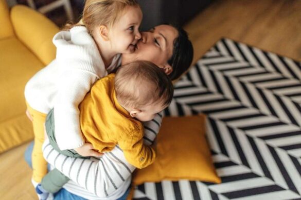 Une mère tient tendrement son bébé et reçoit un bisou de sa petite fille, un moment de complicité familiale avant de voyager avec des enfants.