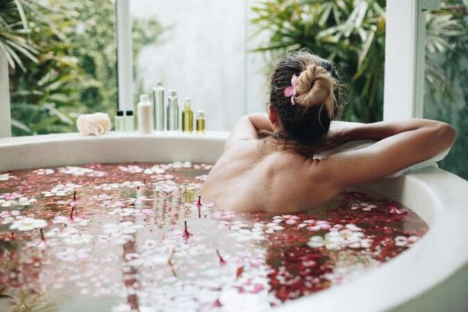 Une femme de dos, cheveux en chignon, se relaxe dans une baignoire remplie d'eau et de pétales de fleurs, avec une vue sur une végétation luxuriante.